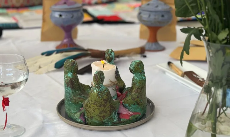 Figuras de cerámica verde en círculo alrededor de una vela blanca, con copas sobre un mantel blanco